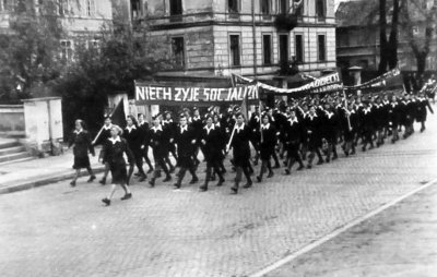 Czarnobiała fotografia przedstawiająca mężczyzn i kobiety idących w pochodzie ulicami miasta.