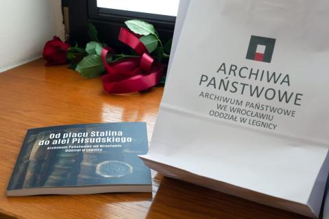 Kwadratowa książka, biała papierowa torba z logiem legnickiego oddziału Archiwum Państwowego we Wrocławiu oraz czerwona róża leżące na drewnianym parapecie okiennym.