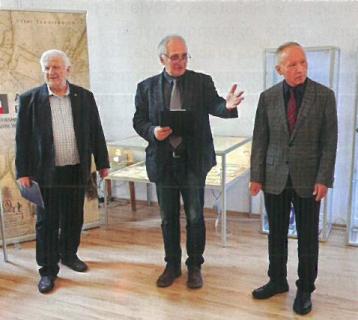 Trzech elegancko ubranych mężczyzn w marynarkach i koszulach stoi na środku sali wystawowej. Za nimi szklana gablota wystawowa i baner.