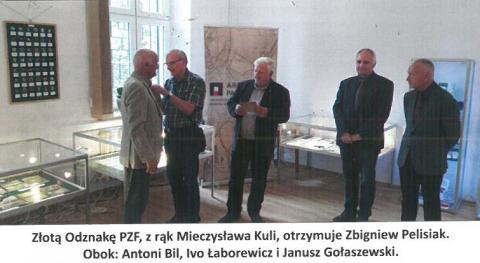 Pięciu elegancko ubranych mężczyzn w marynarkach i koszulach stoi na środku sali wystawowej. Za nimi szklane gabloty wystawowe i baner. Mężczyzna po lewej przypina drugiemu mężczyźnie odznakę.