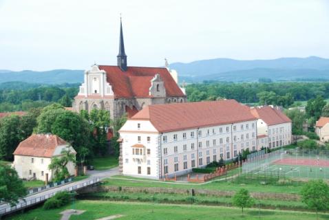 Na pierwszym planie bardzo duży, stary, zabytkowy, czteropiętrowy budynek pokryty pomarańczową dachówką będący siedzibą oddziału Archiwum w Kamieńcu Ząbkowickim. W tle stary kościół oraz góry. 