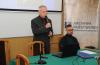 Mężczyzna w garniturze i z mikrofonem w ręku prowadzi wykład w sali konferencyjnej. Po prawej stronie przy stole i przy komputerze siedzi drugi mężczyzna.