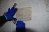 Dwie dłonie w niebieskich, gumowych rękawiczkach podtrzymują rozwinięty na stole stary dokument pisany odręcznie w języku niemieckim.