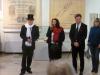 Dwóch mężczyzn i dwie kobiety odświętnie ubrane stoją na tle ekspozycji muzealnej. Mężczyzna z lewej ma na sobie czarny kapelusz i biały szalik oraz garnitur. Kobieta w średnim wieku po środku z mikrofonem w ręku zwraca się do gości.