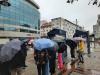 Grupa młodzieży licealnej zgromadzona na placu miejskim z parasolkami w dłoniach stoi na wybrukowanym placu obok wystawy poświęconej Mikołajowi Kopernikowi. Pada deszcz. W tle nowoczesny, przeszklony budynek biurowy.