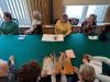 Mężczyźni i kobiety siedzą przy długim stole nakrytym zielonym obrusem w sali konferencyjnej archiwum. Seniorzy słuchają wykładu. Na pierwszym planie trzy osoby, które mają przed sobą stary poszyt akt i oglądają go z zaciekawieniem.