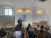 Młody, tęgi mężczyzna w ciemnej marynarce i spodniach oraz w fioletowej koszuli trzyma mikrofon w ręku i przemawia. Stoi na tle podświetlonych szklanych gablot wystawowych, które prezentują stare dokumenty. Jest zwrócony w stronę dzieci i dorosłych, którzy siedzą na krzesłach po środku dużej sali wystawowej. Po lewej stronie widać okno zasłonięte szafą. Zgromadzeni słuchają mężczyzny.