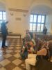 Duża sala wystawowa w stylu barokowym z łukowatym sklepieniem i podłogą w biało-brązowe kafle. Młody, krótkościęty mężczyzna w ciemnej marynarce trzyma prawą rękę uniesioną do góry wskazując sufit. Na krzesłach po środku sali siedzą goście, którzy spoglądają w górę na sufit. W tle duże okna.