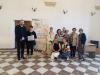 Duża sala wystawowa w stylu barokowym z łukowatym sklepieniem i podłogą w biało-brązowe kafle. Grupa ośmiu starszych kobiet i dwoje archiwistów (młody mężczyzna i kobieta) stoją po środku sali i pozują do wspólnej fotografii. Za nimi stoi baner z logo kamienieckiego oddziału Archiwum Państwowego we Wrocławiu.