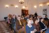 Duża sala wystawowa z dużymi drewnianymi oknami oraz podłogą w biało-brązowe kafle. Po środku sali na drewnianych krzesłach siedzi grupa osób dorosłych i dzieci. Nad nimi świeci duży żyrandol. W tle w rogu sali są otwarte drewniane drzwi.