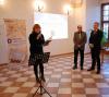Kobieta ubrana na czarno z mikrofonem w ręku trzymająca otwartą teczkę w lewej dłoni oraz dwóch mężczyzn w garniturach po prawej stronie stojących na tle dużego okna. Stoją na środku sali wystawowej na tle ściany z wyświetloną prezentacją multimedialną. Po lewej stronie stoi baner z napisem archiwa państwowe.