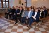 Duża sala wystawowa z dużymi drewnianymi oknami oraz podłogą w biało-brązowe kafle. Po środku sali na drewnianych krzesłach siedzi grupa osób dorosłych, mężczyzn i kobiet. Są elegancko ubrani.