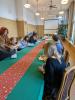 Długa i wąska sala konferencyjna. Po środku długi stół nakryty zielonym obrusem. Grupa studentów siedzi po obu stronach stołu i słuchają wykładu. Niektórzy robią notatki.