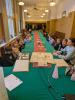 Piętnaście dzieci, nauczyciel i nauczycielka siedzą w sali konferencyjnej przy długim stole nakrytym zielonym obrusem i czerwonym bieżnikiem. Na stole leżą stare dokumenty, pluszak w kształcie kostki oraz drewniane podkładki z pieczęciami.