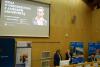 Duża sala konferencyjna w układzie amfiteatralnym. Dwie kobiety prowadzą wykład prezentujący grę planszową. Kobieta po prawej w blond włosach mówi do mikrofonu. Za nimi na dużym ekranie wyświetlona prezentacja zatytułowana "Graj w dziedzictwo z Jamesem Archiwistą". W tle stoją banery z napisem Biblioteka Uniwersytecka we Wrocławiu.