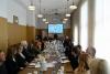 Sala konferencyjna, liczna grupa osób siedzi przy długim stole, a na jej końcu, pośrodku stoi mężczyzna z mikrofonem. Nad nim wyświetla się slajd o konkursie.