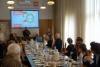 Sala konferencyjna, liczna grupa osób siedzi przy długim stole, a na jej końcu, pośrodku stoi mężczyzna z mikrofonem. Nad nim wyświetla się slajd o konkursie.
