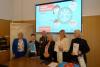 Sala konferencyjna. Sześć elegancko ubranych osób, w tym cztery kobiety, mężczyzna i chłopiec, pozują do zdjęcia uśmiechając się. Dwie kobiety oraz chłopiec trzymają otrzymane dyplomy i torby z upominkami. W tle wyświetla się slajd o konkursie genealogicznym oraz widać plakat ze starą mapą Wrocławia.
