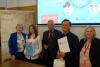 Pięć osób, w tym dwie kobiety, dwóch mężczyzn i dziewczyna, pozują do zdjęcia uśmiechając się. Jeden z mężczyzn oraz dziewczyna trzymają otrzymane dyplomy i torby z upominkami. W tle wyświetla się slajd o konkursie oraz widać plakat ze starą mapą Wrocławia.