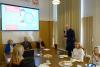 Sala konferencyjna. Na pierwszym planie fragment stołu, po jego prawej stronie stoi wysoki mężczyzna z mikrofonem i gestykuluje. Po lewej stronie dwie kobiety oraz mężczyzna patrzą na przemawiającego z zainteresowaniem.