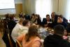 Sala konferencyjna, przy długim stole, po jego obu stronach siedzi liczna grupa studentów. Jedna studentka stoi i gestykuluje, na stole leżą rozłożone gry.
