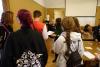 Sala konferencyjna. Archiwistka w blond włosach rozdaje młodym ludziom kartki z zadaniami. Młodzież stoi w kolejce po kartkę z zadaniami. W tle na sali widoczne stoły z rozłożoną grą planszową oraz dorosłych i dzieci grających w grę.