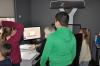 Pracownia digitalizacji. W centrum stanowisko komputerowe, przy którym siedzi pracowniczka i patrzy w ekran monitora. Obok niej stoi skaner. Wokół stoi czworo dzieci i obserwuje pracę kobiety.