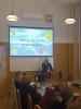 Sala konferencyjna, kobieta stoi z mikrofonem, opowiada i patrzy w stronę grupy siedzących osób. Nad nią wyświetla się slajd z napisem "Emocje są Twoim komentarzem!" i kilkoma emotikonami.