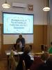 Sala konferencyjna, na ekranie wyświetla się slajd z napisem "Archiwum w akcji! Projektowanie gry planszowej od A do Z", a pod nim stoi kobieta z mikrofonem i opowiada, zwrócona w stronę siedzących przy stole osób.