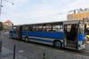Niebieski, zabytkowy, długi, przegubowy autobus, widoczny od boku, stoi na asfaltowej drodze, obok zatoczki dla autobusów. W środku siedzi liczna grupa osób. W tle widoczne są budynki, a w oddali po lewej stronie stoi budynek Archiwum Państwowego we Wrocławiu.