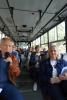 Wnętrze autobusu, obiektyw zwrócony na tył pojazdu od miejsca samego przodu. W pierwszym, najbliższym rzędzie siedzeń, po lewej stronie siedzi starszy mężczyzna i przemawia do mikrofonu, patrząc na obiektyw, a po prawej stronie siedzi pracowniczka o blond włosach. W następnych rzędach siedzi liczna grupa osób, aż do samego tyłu pojazdu.