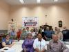 W sali o kremowym kolorze ścian, do zdjęcia pozuje z uśmiechem dziewiętnaście osób. Cztery seniorki i jeden senior siedzą bliżej obiektywu, przy nich stoją stoły, a za stołami stoi młoda kobieta, mężczyzna, osiem seniorek i czterech seniorów. Przy ścianie stoi biała tablica z napisem "Stowarzyszenie Kamieniecki Klub Seniorów".