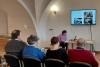 Sala wykładowa z łukowatym sklepieniem. Widoczne plecy gości siedzących i słuchających wykładu. Młoda kobieta w ciemnych włosach średniej długości, różowym swetrze i czarnych spodniach siedzi przy stoliku z laptopem na tle ściany z telewizorem.
