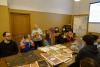 Sala konferencyjna. Na stole rozłożone teczki z dokumentami. Studenci siedzą przy stole. Kobieta w jasnym swetrze i blond włosach trzyma w rękach teczkę aktową i prowadzi zajęcia.