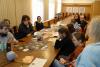Sala konferencyjna. Na stole rozłożone teczki z dokumentami, maskotka i gra planszowa. Studenci siedzą przy stole i przyglądają się grze. Kobieta w jasnym swetrze i blond włosach prowadzi zajęcia.