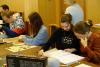 Grupa pięciu studentów i studentek siedzących przy stole. Przeglądają teczki z dokumentami. Za nimi kobieta w blond włosach i niebieskim swetrze nadzoruje ich pracę. Widoczne pudełko z grą planszową na stole.