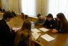 Grupa czterech studentek i student siedzących przy długim stole w sali konferencyjnej. Na stole leży teczka z dokumentami. Wszyscy przeglądają dokumenty leżące na stole.