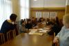 Grupa dziesięciu osób będąca studentami siedzi przy długim stole w sali konferencyjnej. Są podzieleni na dwie grupy. Na stole rozłożone są teczki z dokumentami. Wszyscy przeglądają dokumenty. Dwie kobiety - opiekunka grupy oraz prowadząca wykład stoją i przyglądają się ich pracy.