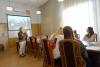 Sala konferencyjna. Kobieta w blond włosach trzyma w dłoniach księgę i pokazuje ją grupie studentek siedzących przy stole.