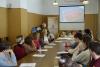 Sala konferencyjna. Grupa czternastu studentek i studentów siedzą przy długim stole. Kobieta pośrodku sali prowadzi wykład. W tle ekran z wyświetloną okładką starej księgi.