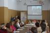 Sala konferencyjna. Grupa studentek i studentów siedzą przy długim stole. Pośrodku kobieta w blond włosach zwrócona jest w stronę ekranu. Wszyscy patrzą na ekran z wyświetlonym starszym mężczyzną z mikrofonem w ręku.