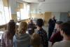 Pracownia konserwatorska. Starszy, łysiejący mężczyzna w ciemnoszarym fartuchu i w okularach stoi naprzeciw grupy ośmiu studentek i studentów i opowiada o pracowni konserwatorskiej. Obok niego stoi profesor.