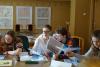 Sala konferencyjna. Cztery studentki siedzą za stołem i przeglądają stare teczki z dokumentami. W tle ściana z widocznymi drzewami genealogicznymi.