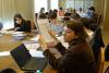 Sala konferencyjna. Przy dwóch stołach siedzą studentki i studenci. Przeglądają dokumenty rozłożone na stole. Pracują w skupieniu. Na pierwszym planie laptop na stole i studentka trzymająca kartki papieru.